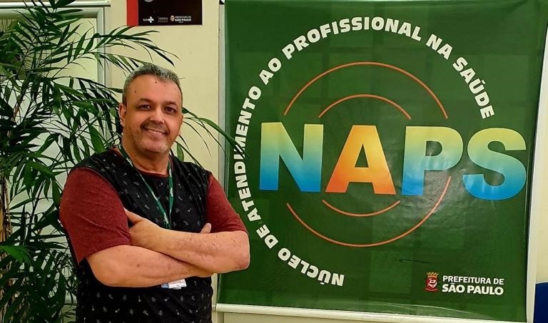 David está sorrindo para a foto e com os braços cruzados. Do lado esquerdo dele há uma planta e do lado direito tem um banner escrito NAPS.